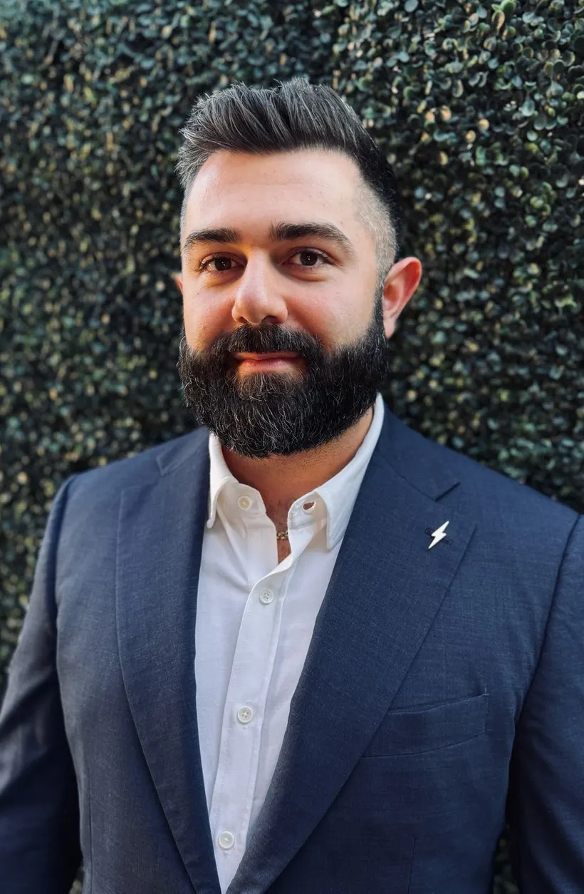 Joshua Palassis in a navy blazer and white shirt, a lightning bolt pin on his lapel